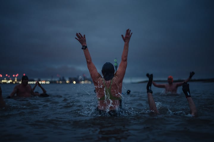 A woman jumps up in the water at dawn. The city of Liverpool is on the horizon.