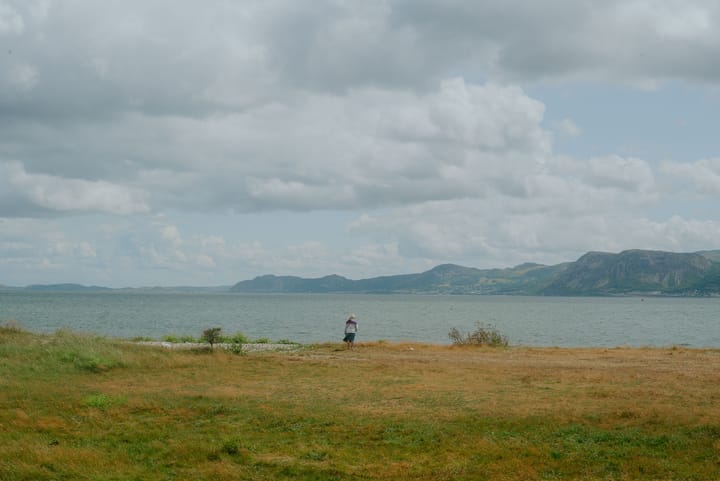 A woman stands alone at the edge of the coast looking to the sea and mountains.