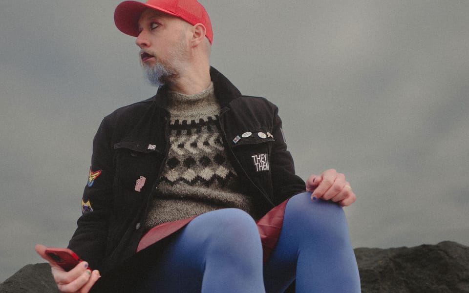 Non-binary person sitting on rocks wearing red skirt, blue tights and denim jacket.